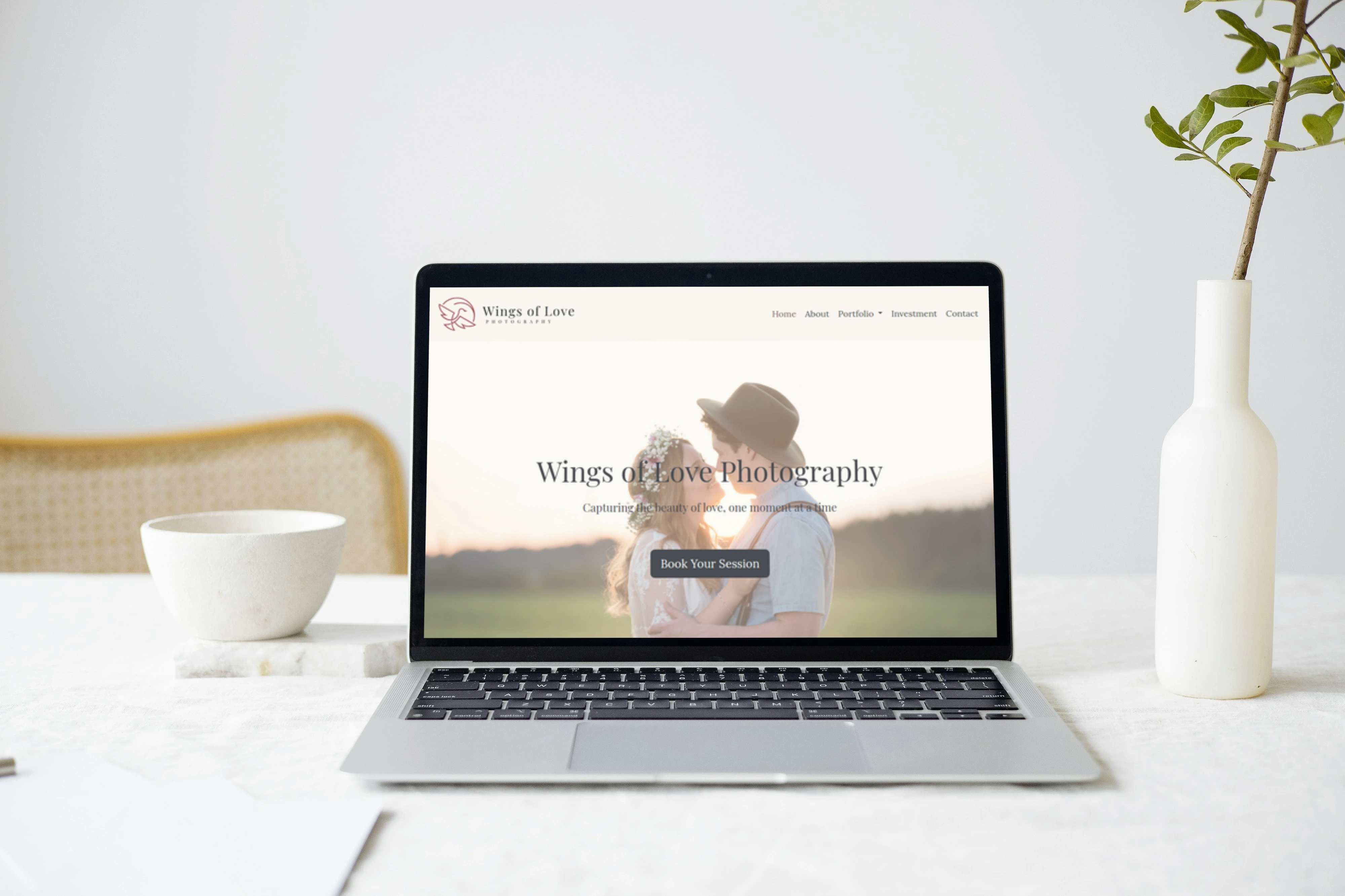 Wings of Love Photography website shown on a laptop in-between a white cup and a white vase on a white table.